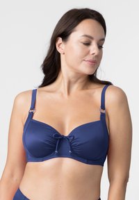 Tamsiai mėlynas bikini viršus su sutrauktu centru, reguliuojamomis petnešėlėmis, metaliniais akcentais ir kaspinu priekyje. Lygus, elastingas audinys.