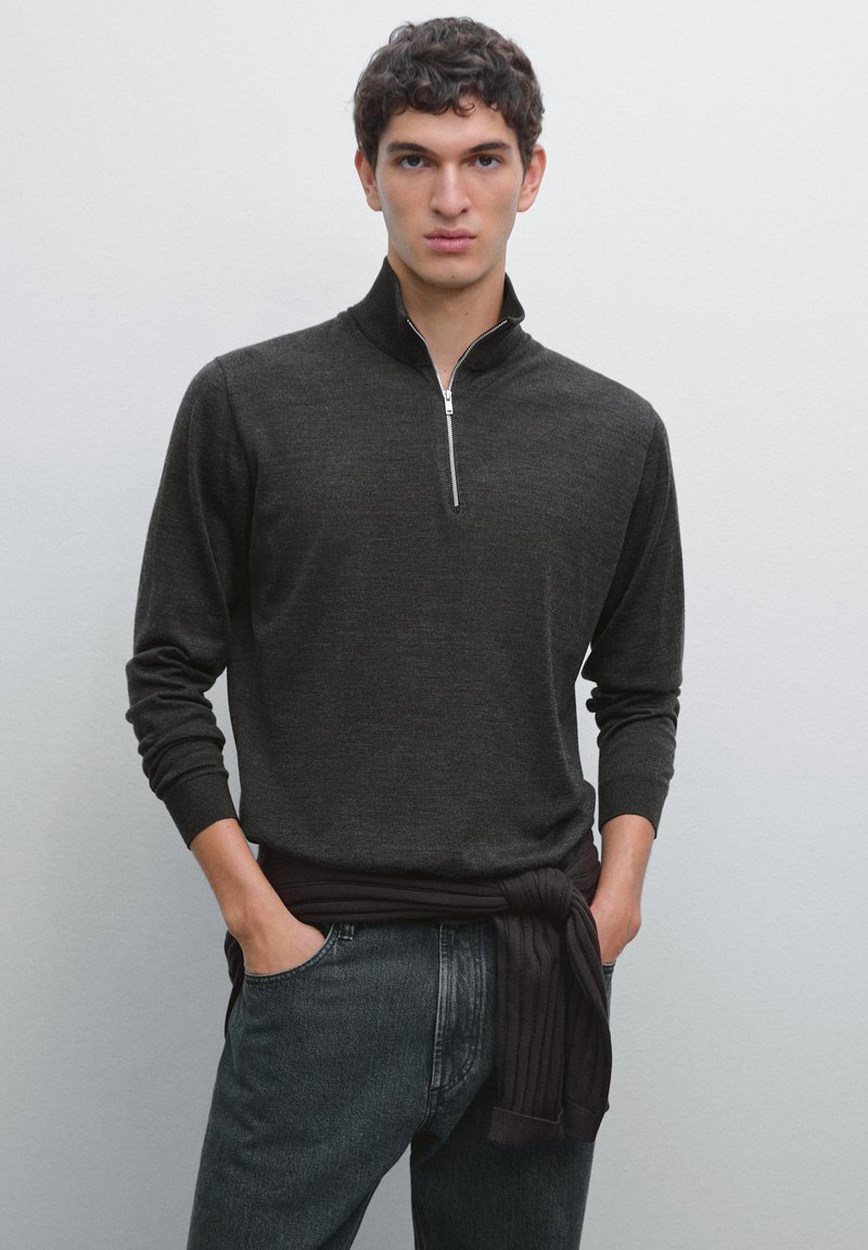 Maglione pullover grigio scuro con colletto a mezza zip, maniche lunghe e polsini a costine, abbinato a jeans di denim scuri e un tessuto scuro legato in vita.
