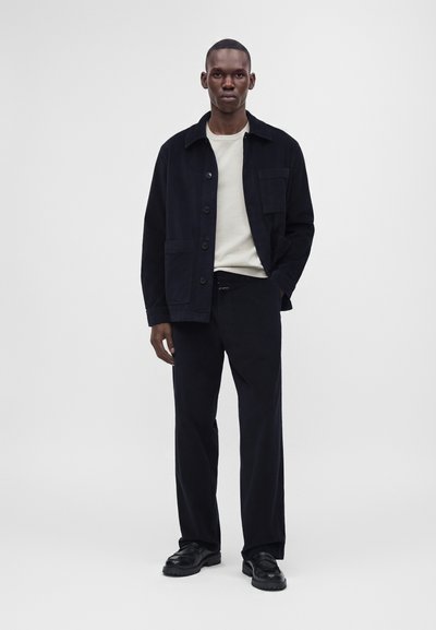 Homme debout portant une veste noire boutonnée, une chemise blanche, un pantalon noir et des chaussures noires sur un fond gris uni.