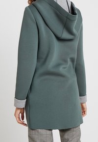 Persoon in een lange, olijfgroene hoodie met grijze manchetten en voering van de capuchon, gecombineerd met zwart-witte geblokte broek, gezien van achteren.