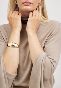 Pronssi ranneketju vaaleanbeige ykkäri päällä, jossa on korkea kaulus ja leveät hihat. Ykkärissä on pehmeä, teksturoitu kangas. Hiukset ovat pitkät ja suorat.