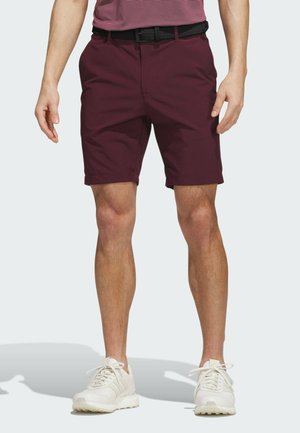Hombre vestido con pantalones cortos rojo oscuro, cinturón negro y zapatillas blancas, de pie frente a un fondo claro con las manos relajadas a los lados.