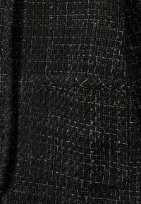 Črn material s teksturiranim vzorcem, ki vsebuje srebrne nitke in vidno žep. Material se zdi tkano in rahlo grobo.