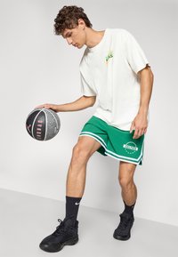 Hvid oversized t-shirt med grafisk print, grønne basketballshorts med hvide striber og "CELTICS" tekst, sorte atletiske sko og struktureret basketball.