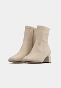 Botine bej până la gleznă, cu toc pătrat, vârf ascuțit și fermoar lateral, expuse pe un fundal alb.