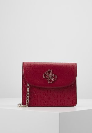 Petit sac bandoulière en cuir rouge avec motif embossé, sangle chaîne argentée et détail logo métallique sur le rabat avant, posé sur une surface blanche.