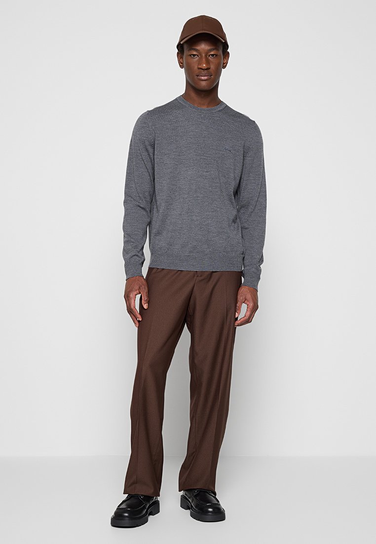 Maglione grigio a maniche lunghe con scollo rotondo, abbinato a pantaloni a righe marroni e scarpe nere. Un cappello marrone completa l'outfit.