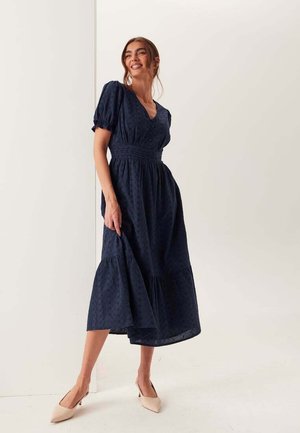 Vrouw glimlachend, draagt een marineblauwe midi-jurk met pofmouwen en een getailleerde taille, gecombineerd met beige puntige hakken, staand tegen een effen achtergrond.