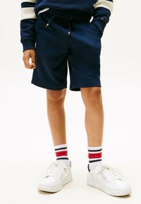 Junge trägt navyblaue Shorts mit Kordelzug, weiße Sneakers und weiße Socken mit roten und schwarzen Streifen, steht vor einem weißen Hintergrund.