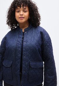 Μπουφάν από τζιν με καπιτονέ σε σκούρο μπλε, με στρογγυλεμένο λαιμόκοψη, κουμπώματα με τρούκ και δύο εμπρόσθιες τσέπες με πατς. Μαλακή υφή με μοτίβο ρόμβων.
