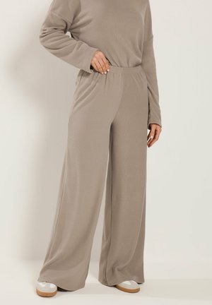 Person iført løstsiddende beige bukser og matchende langærmet top, stående med den ene hånd i bukselommen og hvide sneakers synlige.