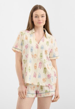 Jeune femme portant une chemise à manches courtes crème à boutons et un short assorti avec des imprimés de chiens colorés sur un fond uni.
