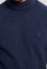 Pull en maille bleu marine avec un col montant, une texture côtelée et un petit logo violet sur le devant, côté gauche.