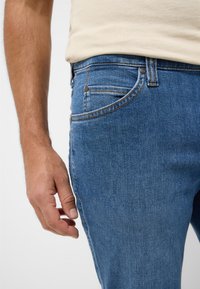 Niebieskie dżinsy o wąskim kroju, z pomarańczowymi stębnowaniami i przednią kieszenią. Ręka spoczywa na boku uda.