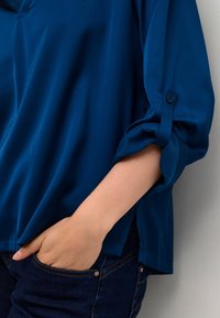 Blouse bleu foncé à coupe décontractée, avec des poignets roulés et une texture douce et lisse ; assortie à un jean foncé avec des détails de poche.