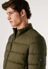 Doudoune vert olive avec un col montant, texture matelassée horizontale, fermeture éclair sur le devant et patch de marque sur la manche gauche.