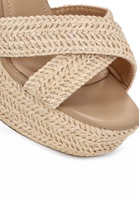 Sandalia de plataforma beige con tiras de paja trenzadas y una zona de dedos de cuero suave, que presenta un diseño redondo de punta abierta y una suela exterior texturizada.