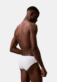 Un slip de bain blanc sur une personne vu de dos, mettant en valeur un design lisse et ajusté avec une taille basse et des lignes épurées sur un fond neutre.