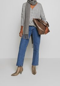 Kobieta w niebieskich dżinsach, szarej zapinanej bluzce, szaliku w kolorze szarym, butach na kostkę z wężowej skóry i trzymająca dużą brązową skórzaną torbę.