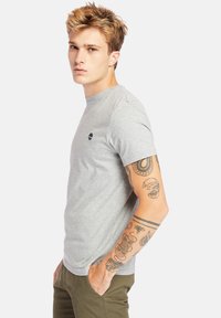 Jonge man met blond haar, gekleed in een grijze t-shirt en olijfgroene broek, met gedetailleerde tattoo's op zijn rechterarm, handen in zijn zakken.