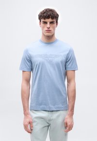 Νεαρός με κατσαρά μαλλιά που φοράει ένα γαλάζιο μπλουζάκι με το λογότυπο της Aston Martin και ανοιχτόχρωμα τζιν, στέκεται μπροστά σε απλό φόντο.