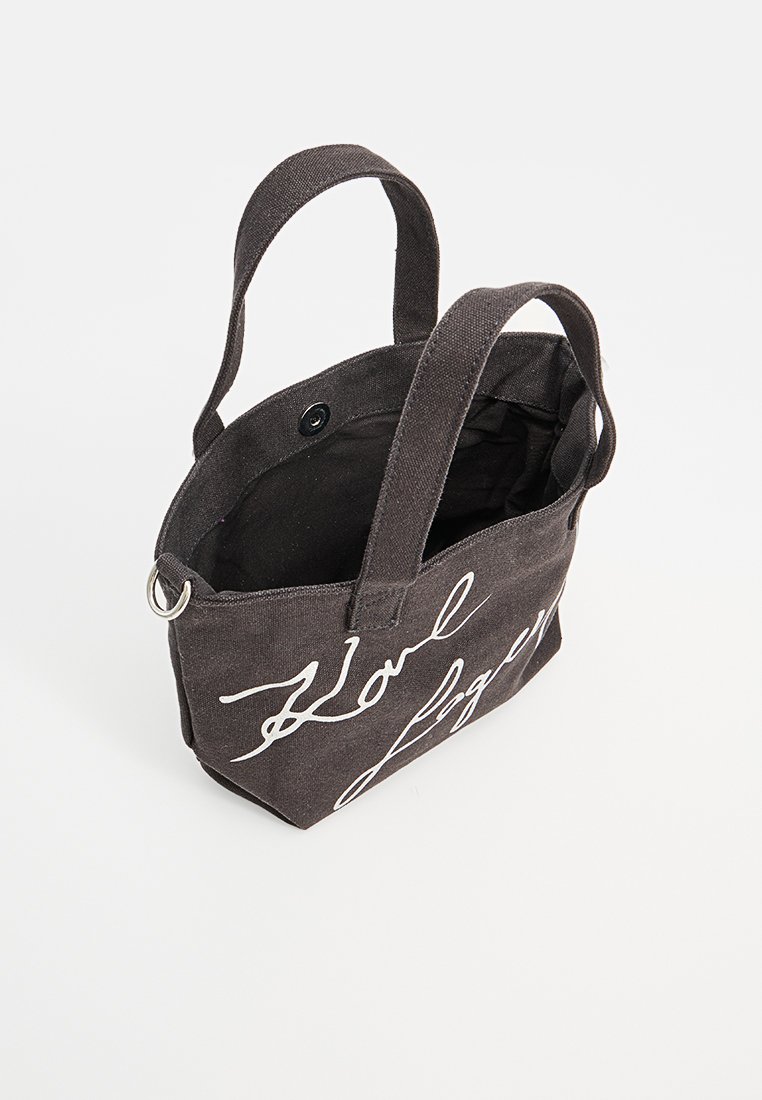Bolsa de lona negra con texto en cursiva blanca y dos asas, que cuenta con un cierre a presión y un detalle de anillo metálico.