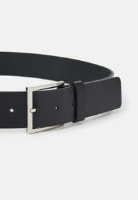 Zwarte leren riem met een rechthoekige zilveren gespsluiting, voorzien van meerdere aanpassingsgaten en een logo-accent nabij de gesp.