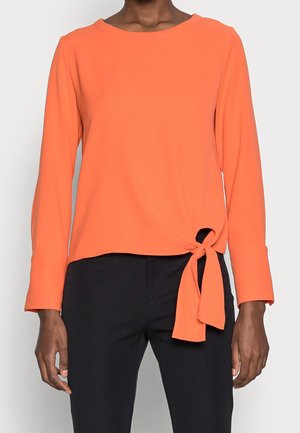 Femme portant un haut orange à manches longues avec un détail noué sur le côté, associé à un pantalon noir, sur un fond clair uni.