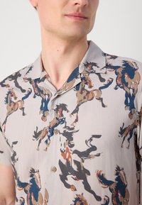 Short-sleeve shirt in beige fabric featuring a repeated horse silhouette pattern in brown, blue, and black. Button detailing visible.