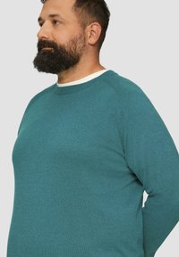 Türkiser Pullover mit Rundhalsausschnitt und gerippten Bündchen, der ein strukturiertes Strickmuster und einen kontrastierenden weißen Innenkragen aufweist.