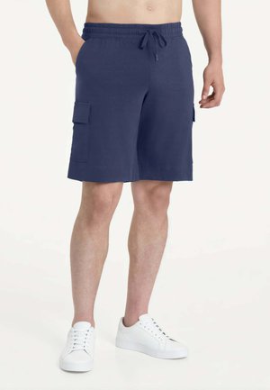 Uomo che indossa pantaloncini cargo blu navy al ginocchio con vita a coulisse e sneakers bianche, in piedi davanti a uno sfondo chiaro e uniforme.