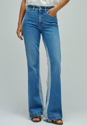 Mujer vistiendo pantalones vaqueros azules de cintura alta con paneles laterales anchos y deshilachados en el borde crudo, combinados con sandalias marrones de tiras y una camisa blanca metida por dentro.