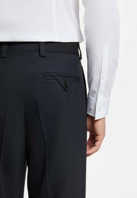 Marineblauwe broek met een gestructureerde stof, voorzien van twee achterzakken met een driehoekige klep en knopdetail. Witte overhemdmanchet zichtbaar.