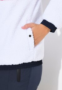 Veste en polaire blanche avec des poignets côtelés navy. Dotée d'une poche latérale avec un bouton-pression noir, mettant en valeur un tissu doux et texturé.