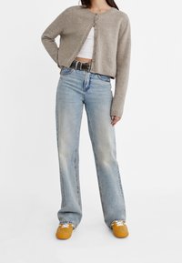 Cardigan corto color marrone chiaro con chiusura frontale a bottoni, indossato sopra un top bianco. Jeans a gamba larga color blu chiaro e scarpe arancioni completano il look.