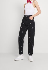 Jeans de mezclilla negros con patrones bordados en forma de corazón en varios colores y un corte de cintura alta. Combinados con zapatillas blancas.