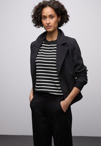 Veste noire cintrée avec grands revers portée sur un pull rayé noir et blanc. Pantalon noir et cheveux courts bouclés. Fond uni.