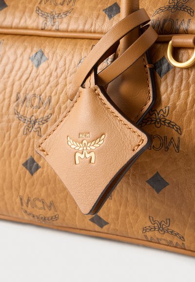 Καφέ τσάντα από δέρμα με υφή, με το λογότυπο και τα σχέδια MCM, με αποσπώμενη ετικέτα με ανάγλυφα χρυσά στοιχεία και διακοσμητικό ράψιμο.