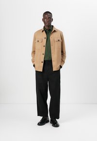 Veste en daim beige avec deux poches poitrine, assortie à une chemise verte foncée et un pantalon large noir. Des chaussures noires complètent la tenue.