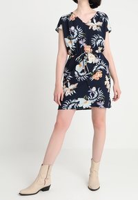 Květinové šaty s krátkým rukávem v námořnické modři, se stahovacími šňůrkami v pase, zdobené barevnými květinami a V-výstřihem. V kombinaci s béžovými kotníkovými botami.
