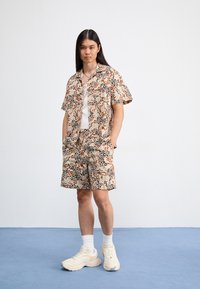 Camicetta a maniche corte e pantaloncini abbinati in tessuto beige con stampa animale astratta in marrone, nero e arancione. Sneakers bianche e calze.