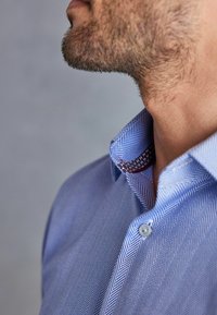 Blauw overhemd met een herringbonepatroon en een button-down kraag. De binnenste kraag heeft een contrasterend rood en wit ruitpatroon.