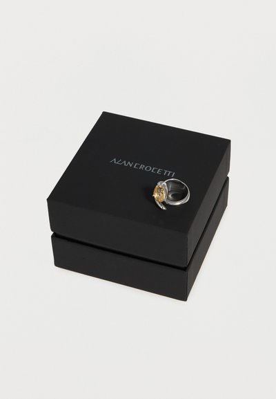 Ασημένιο δαχτυλίδι με χρυσή οβάλ λεπτομέρεια που βρίσκεται πάνω σε μια κλειστή μαύρη κοσμηματοθήκη με το όνομα της μάρκας στο καπάκι.