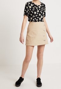 Svart blommig blus med gula och vita prästkragar, parat med en beige A-linjeformad kjol med en framficka och knappdetaljer. Svarta sneakers.