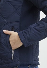 Jachetă capitonată de culoare albastru închis, cu cusături diagonale, buzunar lateral cu fermoar și panouri laterale elastice pentru un confort și o potrivire îmbunătățite.