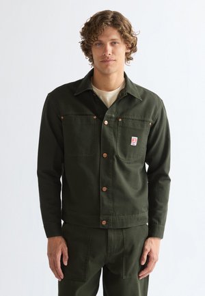 Mann mit lockigem Haar, trägt eine dunkelgrüne, geknöpfte Arbeitsjacke und passende Hose vor weißem Hintergrund.