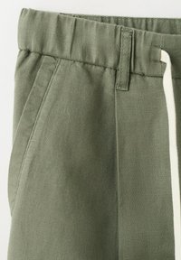 Legere Hose in Olivgrün mit elastischem Bund, Seitentasche, Gürtelschlaufen und weißem Kordelzugdetail auf der rechten Seite.