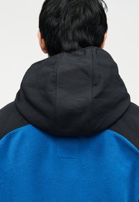 Άτομο με κοντά μαύρα μαλλιά που φοράει μπλε μπουφάν με μεγάλο μαύρο κουκούλι, φαίνεται από πίσω σε απλό φόντο.