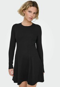 Młoda kobieta z krótkimi blond włosami, ubrana w dopasowaną czarną sukienkę z długim rękawem, duże kołowe kolczyki i złoty pierścionek na lewej dłoni.