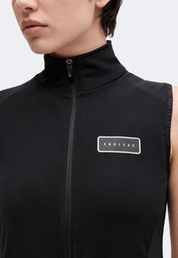 Gros plan d'une personne portant un haut noir sans manches zippé avec un col haut et un logo blanc "ENDLESS" sur la poitrine.